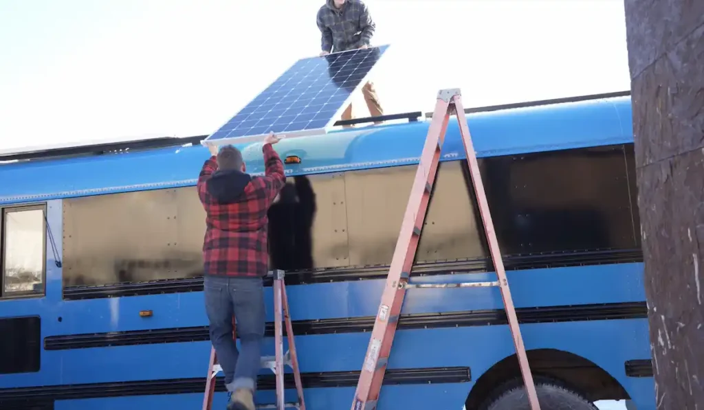 Two people installing solar panels on a skoolie bus roof during an off-grid solar conversion